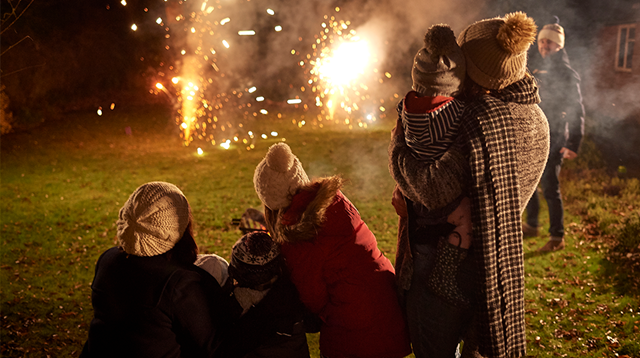 Two young families standing outside in garden on cold, dark night, watching fireworks together